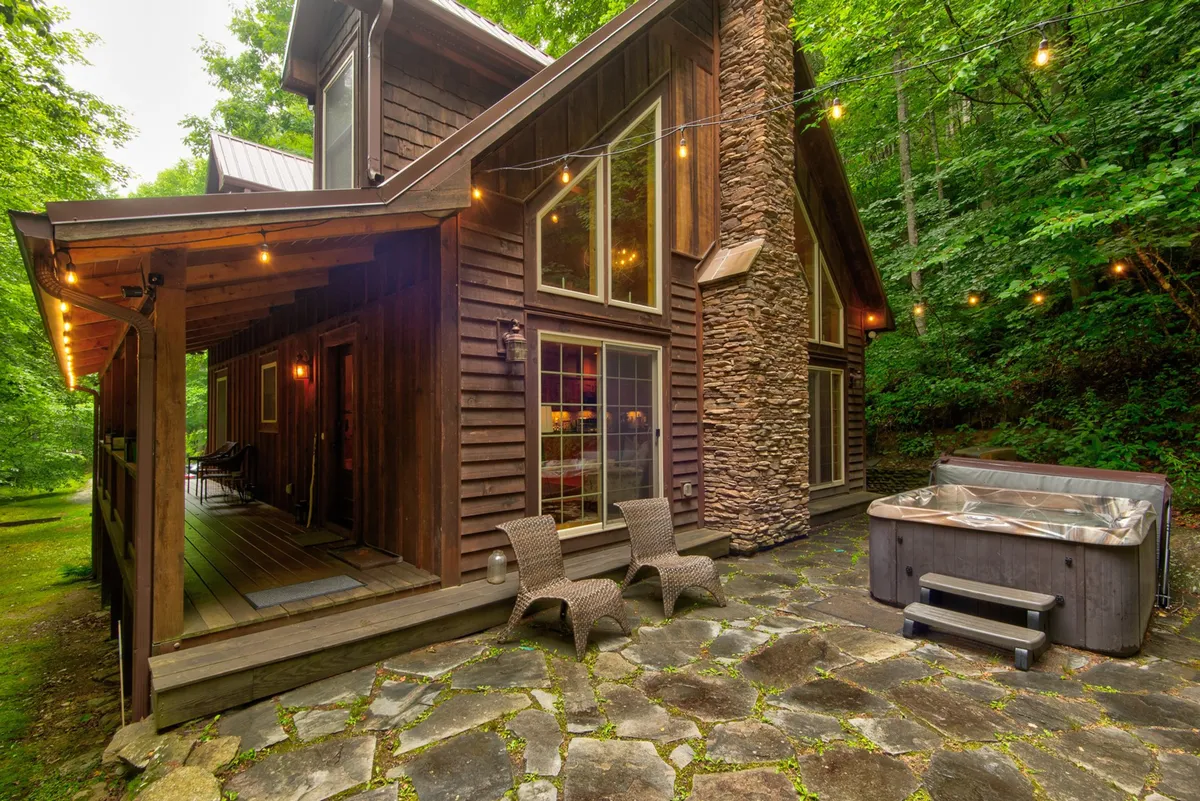 This exterior image features a stone wall, a hot tub, and an inviting outdoor seating area strung with cozy lights. The hot tub is positioned to enjoy serene sunset views, while the surrounding space is perfect for relaxing after a day of winter sports.