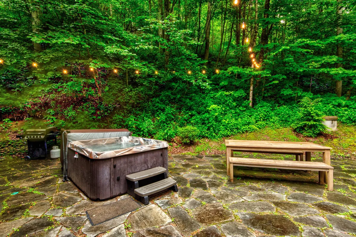 This image features an inviting outdoor space with a hot tub, wooden picnic table, and string lights. The scene radiates warmth and encourages relaxation, with space for alfresco meals and entertaining family or friends. The setup suggests a cozy evening under the stars or fun daytime activities.