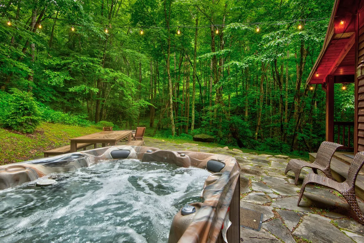 A serene hot tub set on a wooden deck, surrounded by woods and glowing with twinkle string lights. Outdoor seating and a peaceful, inviting mood make this a perfect spot for relaxation and indulgence.
