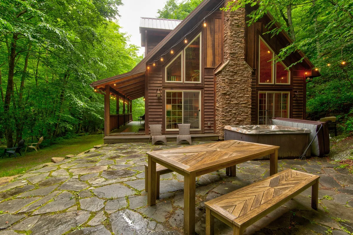 This inviting patio scene includes a wooden table with benches, a hot tub, stone walls, and surrounding greenery, all under string lights. The layout encourages outdoor dining, relaxation, and social connection in a picturesque natural setting.