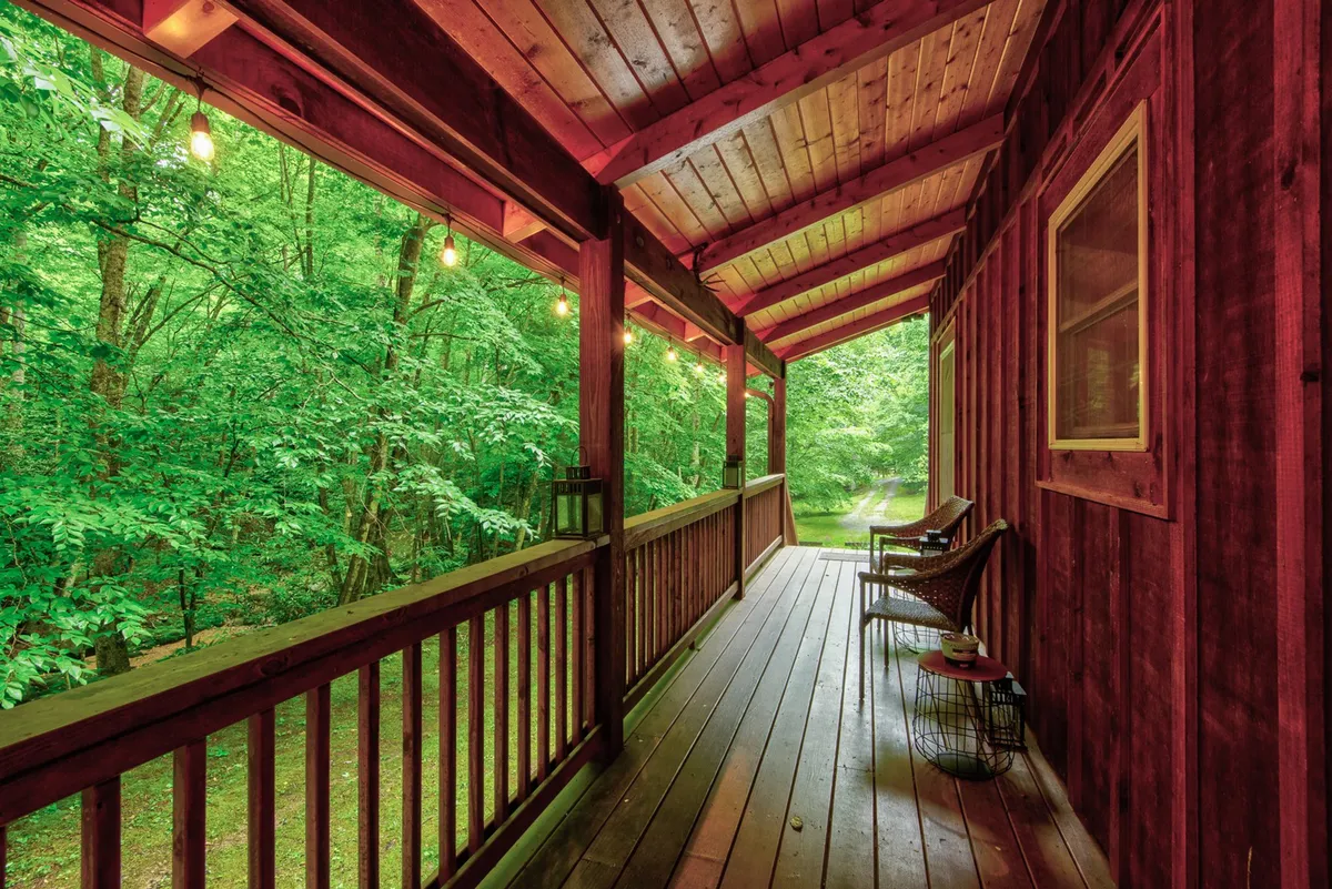 This image captures a long, wooden-covered front porch with classic chairs, hanging lights, a wooden railing, and lush greenery surrounding the area. The porch is inviting and exudes a peaceful, relaxing atmosphere—ideal for enjoying morning coffee or winding down in the evening.