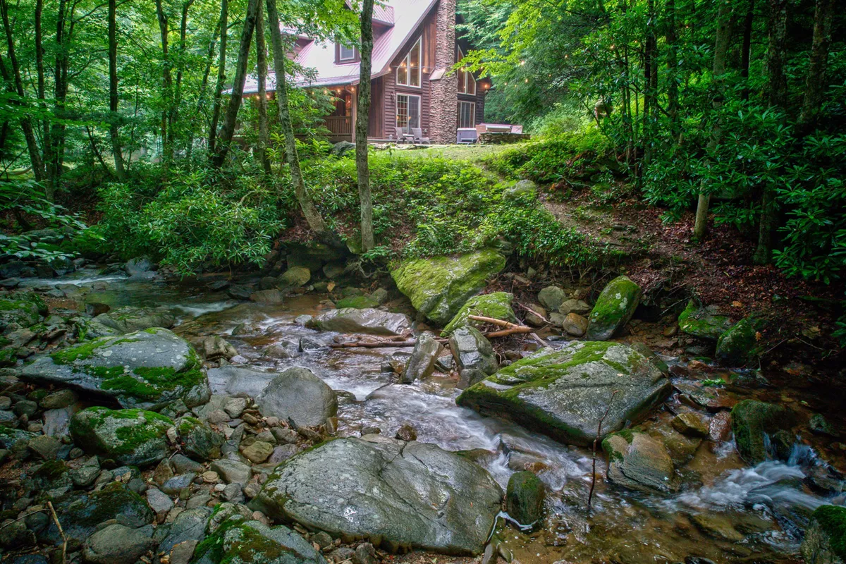 The image showcases a tranquil stream bordered by rocks and trees, with the house nestled nearby. It evokes a sense of adventure and connection with nature, inviting families to explore, fish, or simply relax by the water's edge.