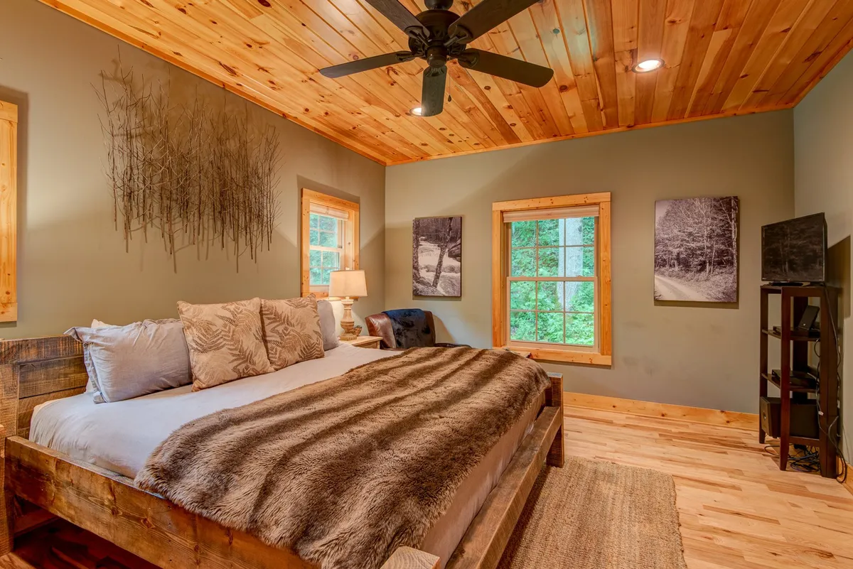 A spacious bedroom featuring a large wooden bed dressed in plush bedding and a faux fur blanket, with wall art above and a large window that lets in abundant natural light and offers a peaceful view of surrounding greenery. The tranquil layout is complemented by an inviting sense of privacy and relaxation, perfect for an evening retreat.