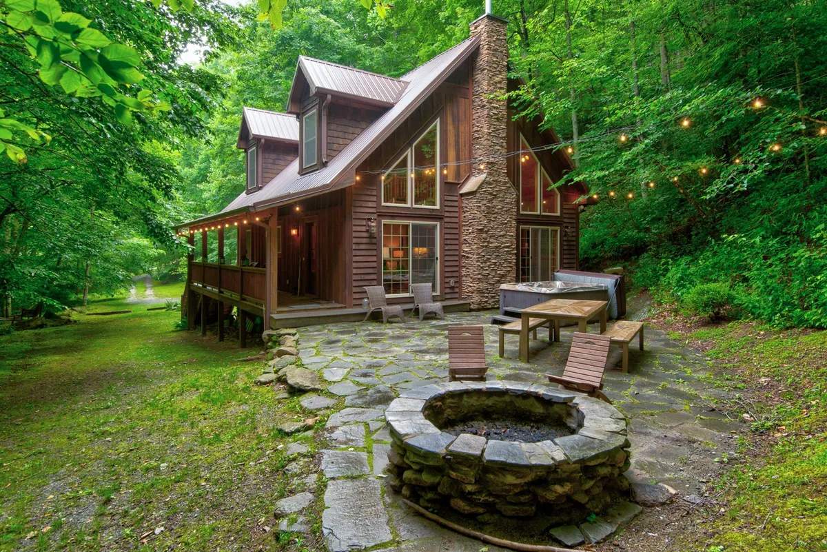 An exterior shot featuring the cabin's distinctive wooden deck, large windows, and a welcoming stone fire pit set amid natural surroundings. The scene captures both architectural charm and key gathering spaces for socializing or stargazing after a day outdoors.