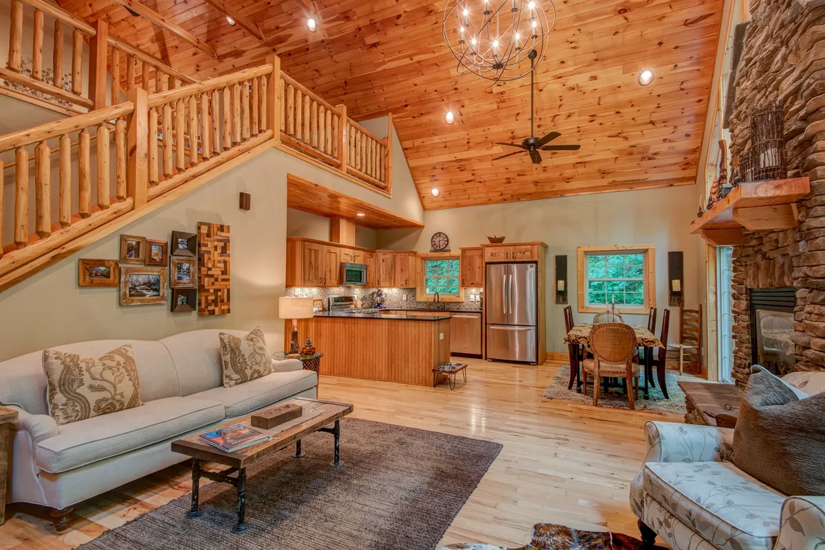 A wide view of the great room, showcasing a plush sofa, a central stacked stone fireplace, a visible wooden staircase, and an open kitchen area. The space is illuminated by ample natural light streaming through large windows, with the dining table for six clearly in view. The rich hickory hardwood floors and vaulted ceilings create an expansive, airy feel.