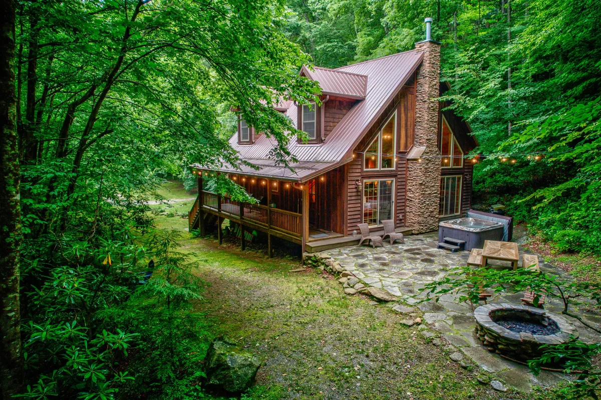 This exterior image showcases the Beech Creek Cabin surrounded by lush greenery, with the hot tub and fire pit visible in the outdoor space. The scene conveys a sense of cozy seclusion, and highlights the amenities available for relaxation after exploring the outdoors.