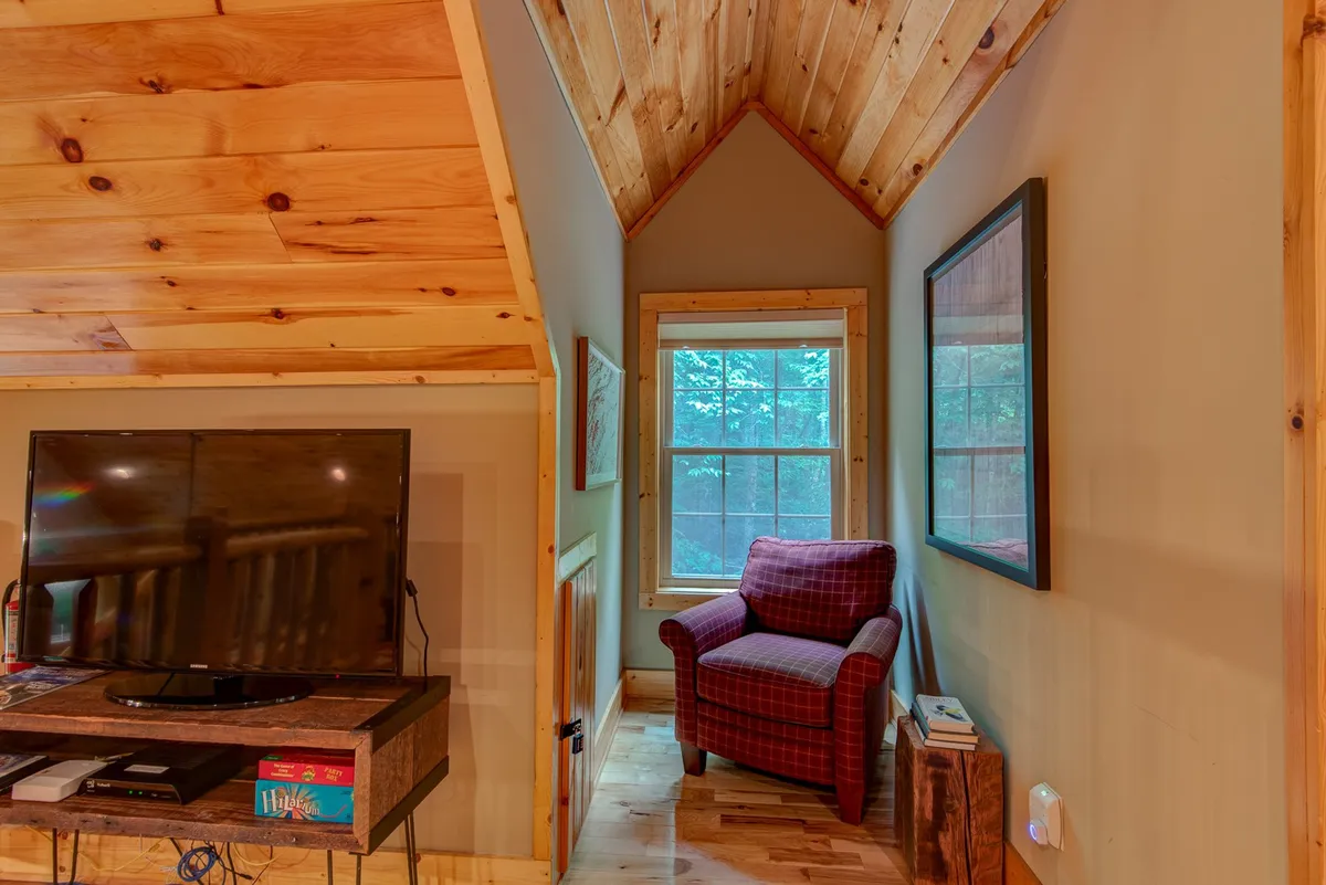 A cozy corner with a Smart TV, a plaid armchair for comfortable viewing or reading, a window letting in natural light, and a wooden ceiling, capturing the inviting and modern aspects of the cabin's living spaces.