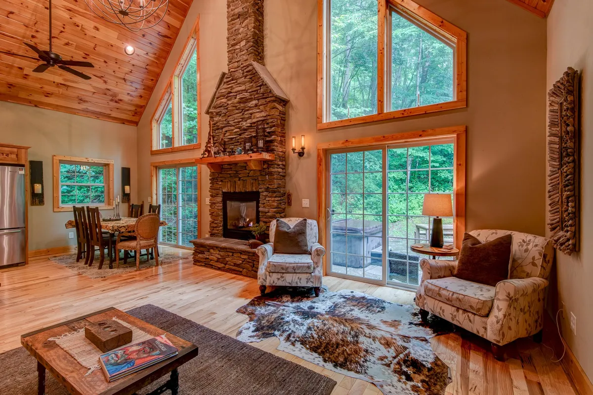 A welcoming view of the great room with a dramatic stacked stone fireplace, comfortable seating arranged for conversation, and expansive windows that frame the forest scenery outside. The adjacent dining area is visible, reinforcing the open-concept design. Sunlight pours in, illuminating the inviting atmosphere.